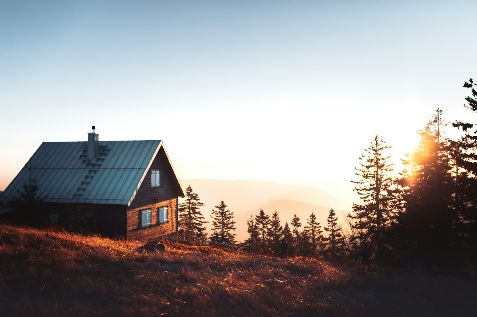 A small wooden cabin on a grassy hill at sunset, with evergreen trees beyond