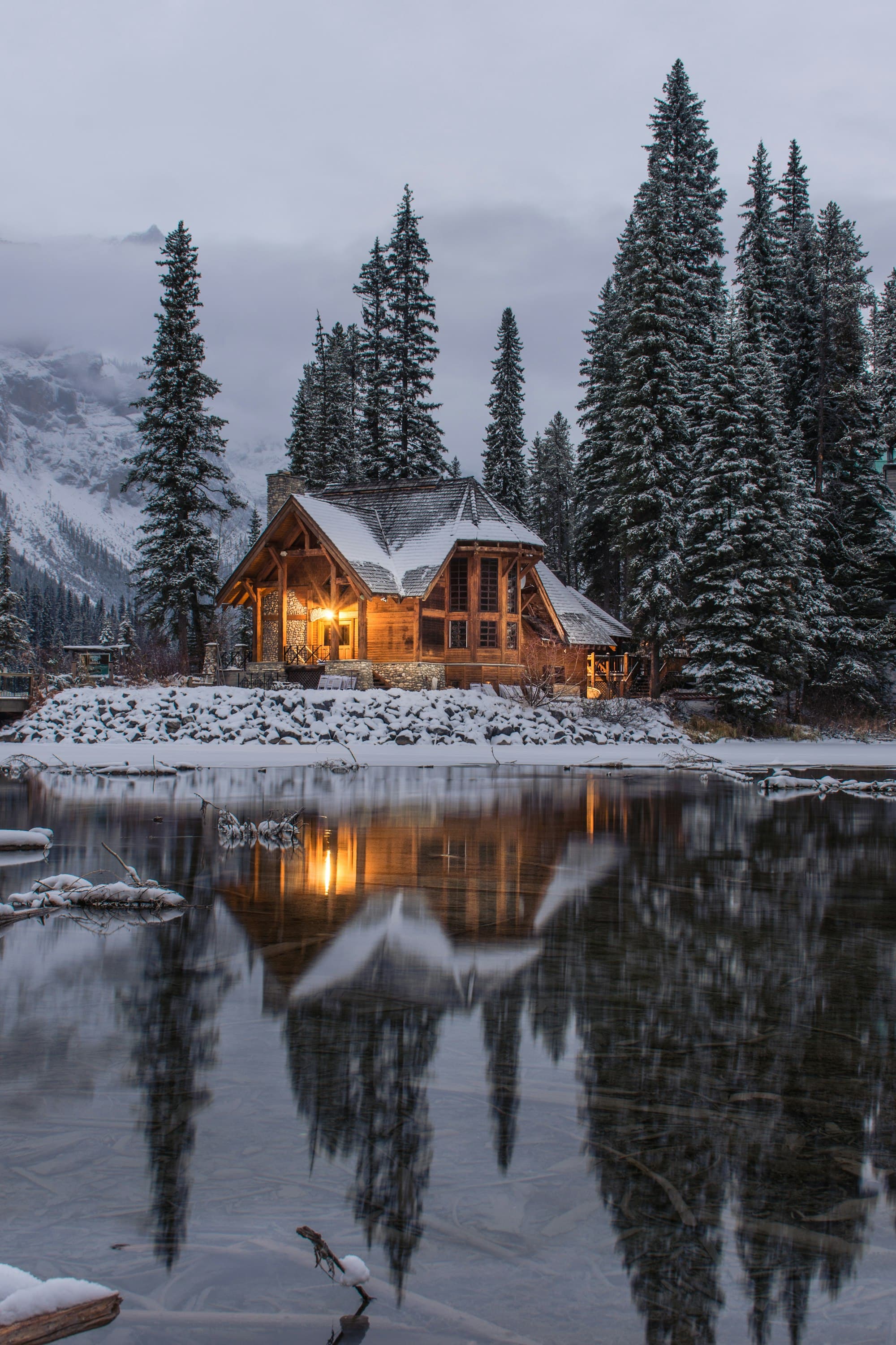 A timber cabin with warm interior light beside a still lake in winter